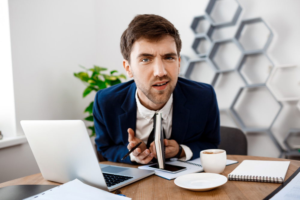 Displeased young businessman sitting at workplace, office background. 